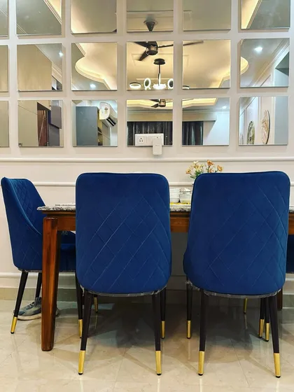 A closer view of the dining set, showing the quilted detailing on the blue velvet chairs and the elegant gold-tipped legs. These details, combined with the reflective quality of the mirror wall, contribute to the room's luxurious and contemporary feel.