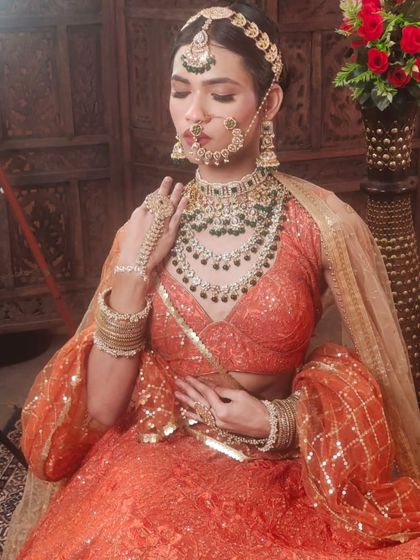 A bride seated in a traditional setting, wearing a heavy, layered green Kundan set with an orange lehenga. The hathphool and maang tikka are beautifully detailed.