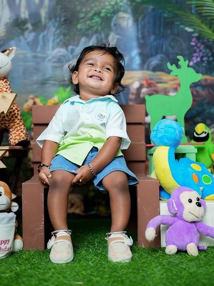A big, happy grin from the birthday boy. The colorful jungle backdrop and cute animal props create a fun and festive atmosphere for a first birthday photoshoot.