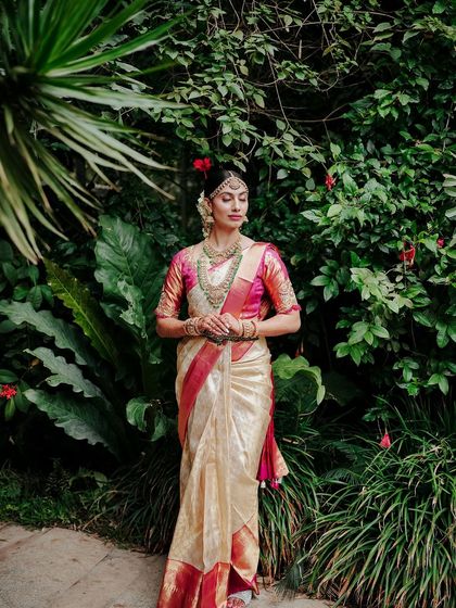 A bride in a traditional South Indian saree stands against a backdrop of lush green leaves. The vibrant colors of her attire pop against the natural setting.