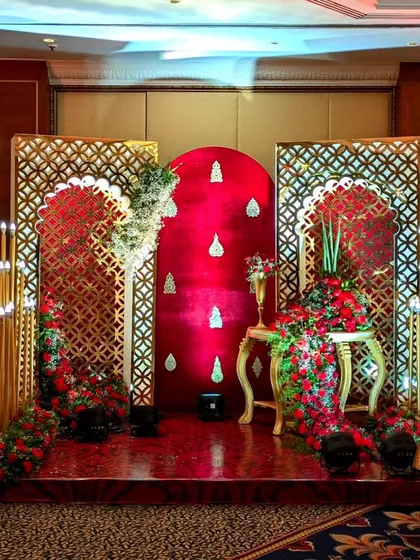 A regal photo corner for a reception, featuring a rich red velvet backdrop, ornate golden lattice screens, and cascading red floral arrangements.