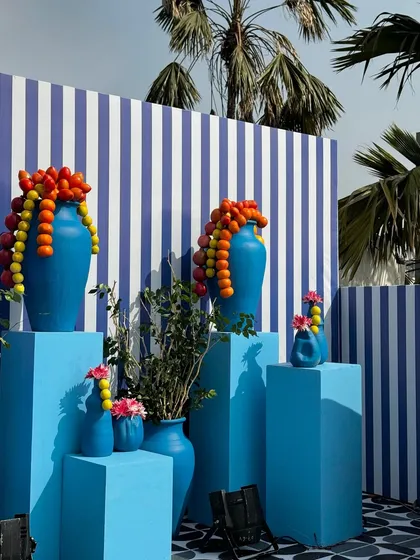 An artistic installation of blue vases on matching pedestals. Instead of flowers, we used garlands of pomegranates and lemons cascading from the vases, creating a unique and colorful focal point against a striped backdrop.