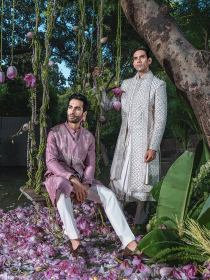This image captures two complementary looks against a lush, natural backdrop. The intricate white sherwani and the elegant mauve kurta set demonstrate our versatility in dressing the modern groom and his closest companions.