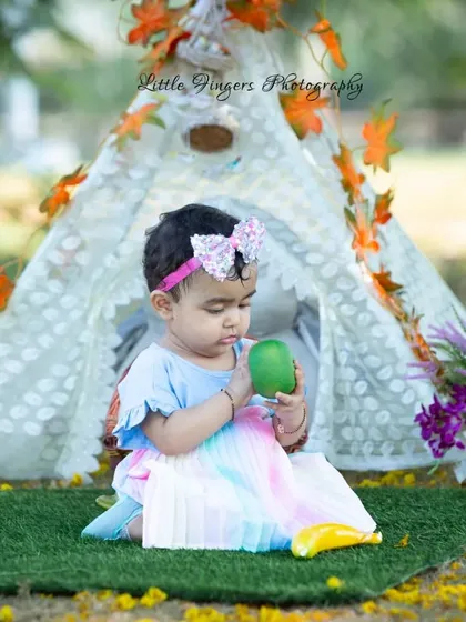 A one-year-old girl curiously examining a piece of fruit in front of her birthday teepee.