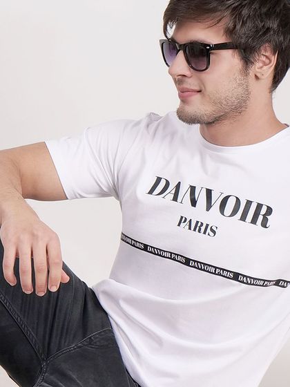 A relaxed, seated pose of a model wearing a white "Danvoir Paris" t-shirt, creating a casual and stylish look.