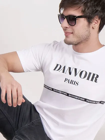 A relaxed, seated pose of a model wearing a white "Danvoir Paris" t-shirt, creating a casual and stylish look.