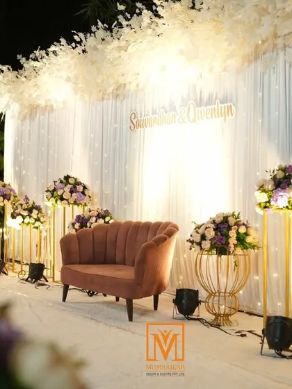 A chic and modern stage setup for a reception or engagement. The backdrop features white drapes and twinkling lights, with a stylish pink sofa and gold floral stands for a touch of glamour.