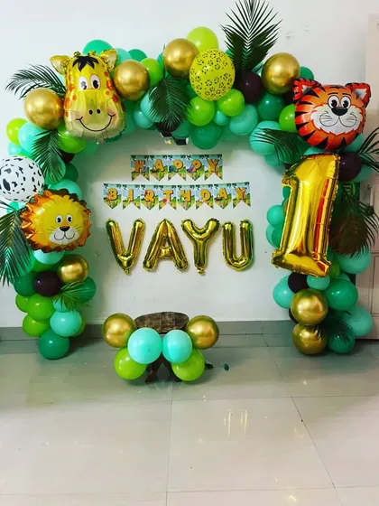 A simple and charming jungle theme balloon arch for a first birthday at home. The setup includes gold letter balloons for the name "VAYU" and cute animal foil balloons.