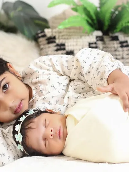 A beautiful portrait of sisterly love. The older sister lies protectively beside her sleeping newborn sibling, looking right at the camera, creating a sweet and engaging photo.