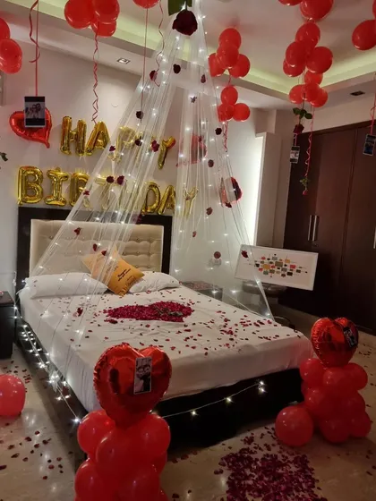 A romantic birthday setup featuring a canopy of fairy lights over the bed, red balloons, rose petals, and hanging photos for a personal touch.