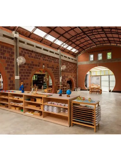 The classroom is filled with natural light from the vaulted ceiling's skylights. The space is organized with low, accessible wooden shelves and learning materials, all set against the warm backdrop of exposed hollow clay block walls.