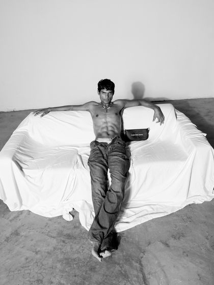 A relaxed, full-body pose from the Calvin Klein series. This black and white shot captures a casual confidence, with the styling focused on a shirtless look paired with jeans and a branded bag.
