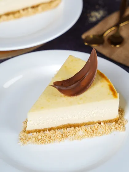 A classic slice of New York cheesecake with a simple chocolate garnish. The clean, minimalist styling on a white plate highlights the creamy texture and perfect bake of the dessert.