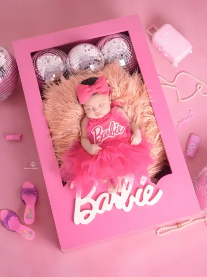 A close-up of a sleeping newborn styled for a 'Barbie' themed shoot. She's dressed in a frilly pink outfit inside a custom Barbie box, showcasing the detailed props I use for themed newborn photography.