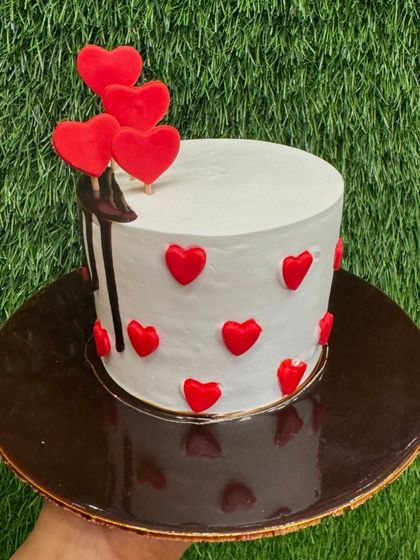 A simple yet striking mini cake with red hearts and a chocolate drip. The contrast between the white frosting, dark chocolate, and red hearts creates a modern and romantic vibe.