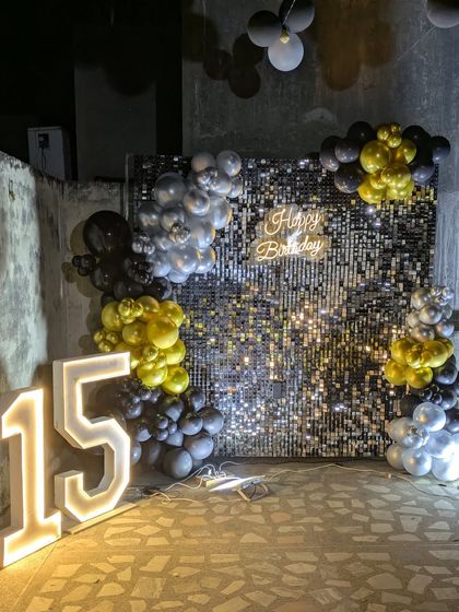 A complete view of the 15th birthday setup with a black sequin wall, gold and silver balloons, and large illuminated numbers.