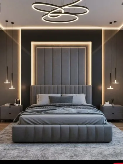 This master bedroom design creates a dramatic and luxurious feel with a floor-to-ceiling upholstered headboard, highlighted by recessed LED backlighting. The layered lighting scheme, including a modern chandelier and hanging bedside pendants, adds depth and sophistication to the contemporary grey palette.
