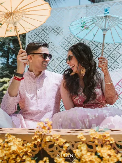A fun and candid moment from a Mehendi or Haldi event. The couple enjoys the celebration with colorful paper umbrellas, seated in a custom-designed space with intricate details.