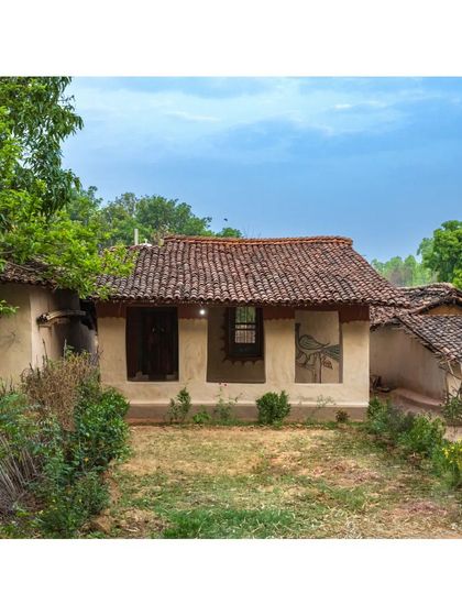 A completed homestay nestled amongst the trees, with its tiled roof and earthen walls blending seamlessly into the rural Madhya Pradesh landscape. The design respects the scale and character of the village.
