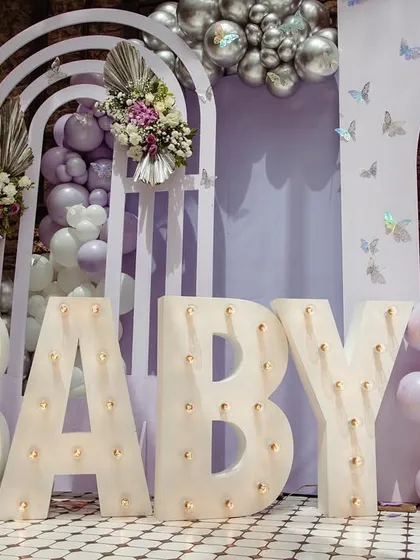 Marquee 'BABY' letters add a warm glow to this lavender-themed baby shower, complemented by balloon arches and floral details.