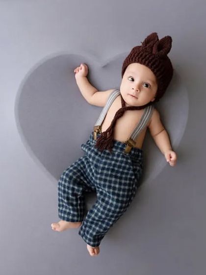 A baby boy in a cute outfit with suspenders and a bunny-ear hat lies on a grey heart-shaped cushion.