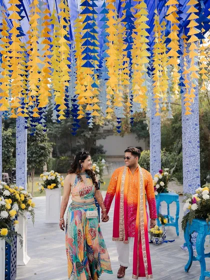 The couple walks under a ceiling of cascading blue and yellow paper tassels, a creative and colorful element that adds texture and movement to the Haldi decor.