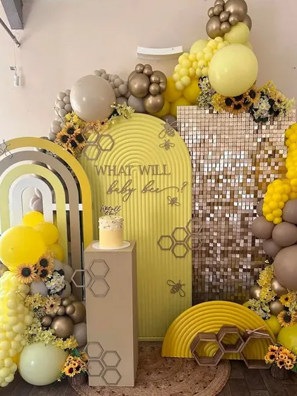 For this unique baby shower, the theme was "What will baby bee?". We designed a modern setup with a shimmer wall, custom arched backdrops, and a beautiful organic balloon garland in shades of yellow, grey, and gold, accented with sunflowers.