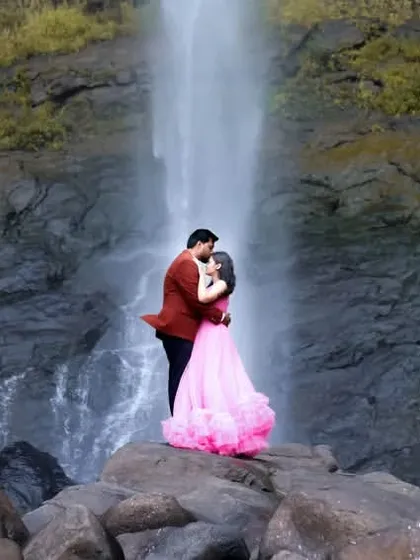 A breathtaking pre-wedding portrait of a couple standing on rocks before a cascading waterfall. The bride's flowing pink gown adds a touch of fairy-tale romance to the dramatic natural landscape.