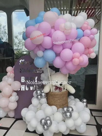 A hot air balloon concept for a baby shower, using a basket with a teddy bear and a large cloud of pink, blue, and white balloons.