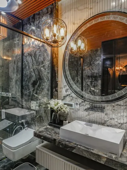 A luxury bathroom in our Vikaspuri project, featuring floor-to-ceiling marble, a large circular mirror with an ornate frame, and a glass-enclosed shower. A classic chandelier adds a touch of opulence.