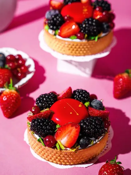 Two exquisite berry tarts on mini pedestals against a solid pink background. The clean, minimalist styling puts the focus entirely on the artistry and fresh ingredients of the dessert.