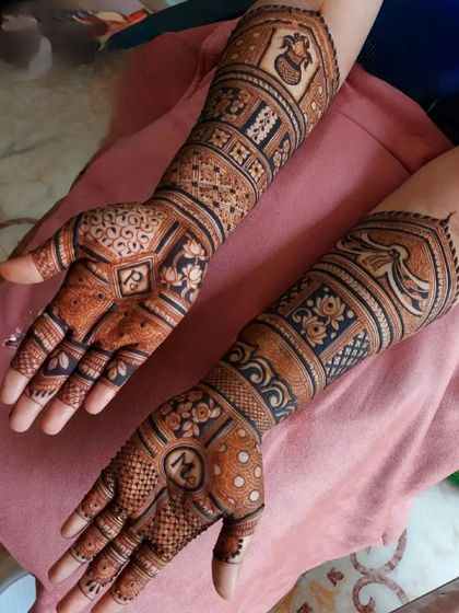 A detailed bridal mehandi with a mix of patterns. One arm features a kalash and floral designs, while the other has geometric checks and traditional figures, personalized with an initial.