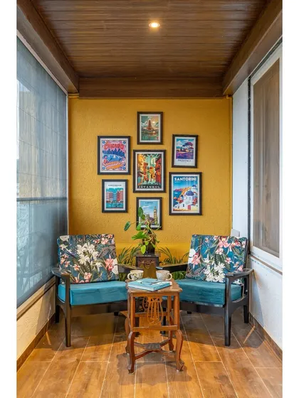 A cozy balcony retreat designed for relaxation. The mustard yellow accent wall, travel-themed gallery, and comfortable chairs with floral cushions make this the perfect spot to unwind.