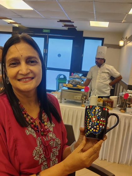 A smiling guest holds up her completed mug at the Sakhi group art party, with the event setup visible in the background.