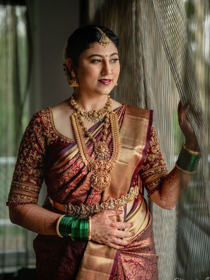 A bride looks out a window, her maroon saree and gold jewelry glowing in the soft light. This is a timeless and elegant bridal portrait.
