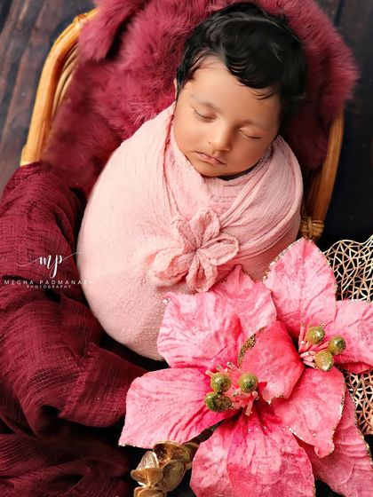 A baby rests peacefully in a cane basket, wrapped in pink and surrounded by rich maroon fabric and a large pink flower.