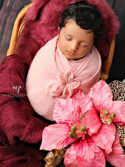 A baby rests peacefully in a cane basket, wrapped in pink and surrounded by rich maroon fabric and a large pink flower.