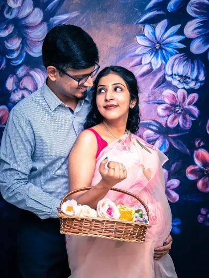 A playful and sweet studio portrait. The couple stands before a floral backdrop, with the mom-to-be holding a basket of flowers.