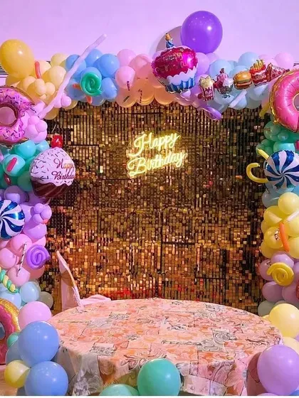 A playful and colorful candyland-themed birthday party. The backdrop features a gold sequin wall, a neon sign, and a balloon arch decorated with donut and cupcake foil balloons.
