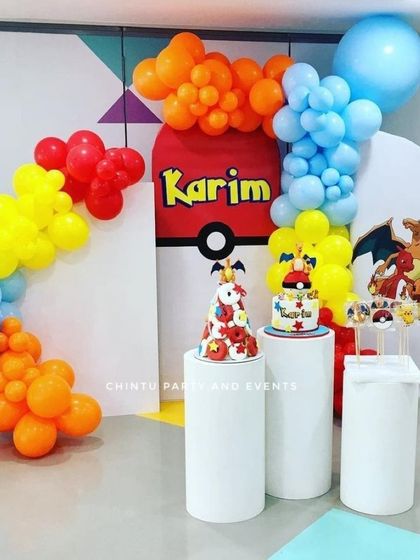 A bright and colorful Pokemon themed birthday decoration. The setup features a balloon arch in primary colors and pedestals for displaying cakes and treats.