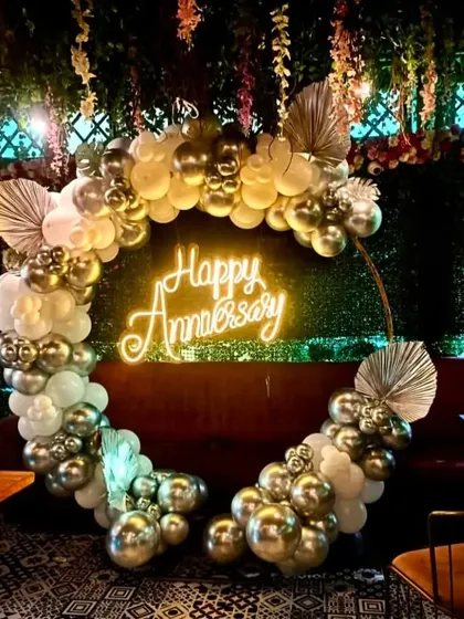 A chic anniversary decoration in a restaurant. The circular balloon garland in white and chrome gold is accented with dried palm leaves and a 'Happy Anniversary' neon sign for a trendy, bohemian look.
