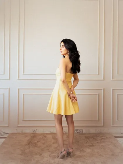 An elegant, full-length shot against a classic paneled wall. The model's pose, looking over her shoulder, showcases the back and silhouette of the yellow strapless dress.