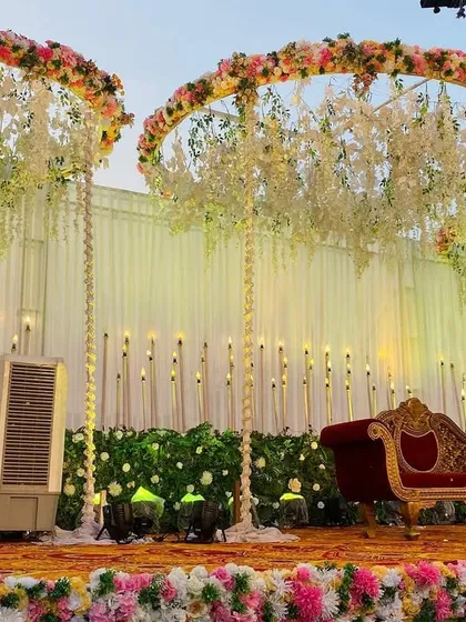 An elegant reception stage with three large, suspended floral rings and a backdrop of warm lights. The red velvet sofa adds a classic touch.