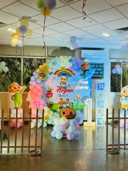 A full Cocomelon themed party setup in a large hall. This decoration includes a central balloon arch, multiple character cutouts on balloon pillars, and a small fence for a playful touch.