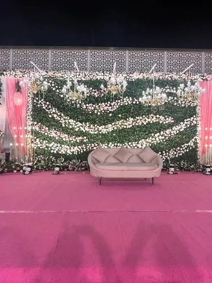 A wide view of a reception stage with a lush green and white flower wall. The pink carpet and hanging globe lights add a modern and romantic touch.