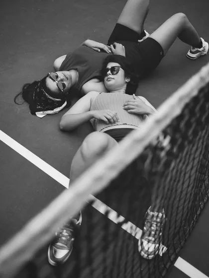 A unique black and white shot from a high angle, showing two women in sunglasses lying on a tennis court, viewed through the net. This image demonstrates creative composition and storytelling in a lifestyle context.