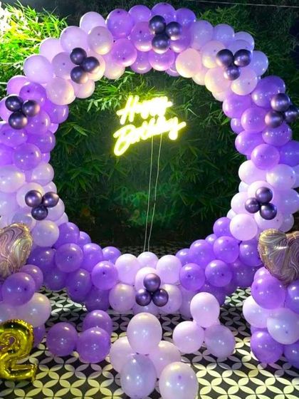 An outdoor birthday decoration featuring a balloon ring in shades of purple. The setup is placed against natural greenery and includes a 'Happy Birthday' neon sign and a number '22' foil balloon.