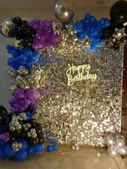 For a modern and glamorous party vibe, I offer sequin backdrops that sparkle. This birthday setup combines a shimmering silver sequin wall with a balloon garland in blue, purple, silver, and black, all brought together with a bright "Happy Birthday" neon sign.