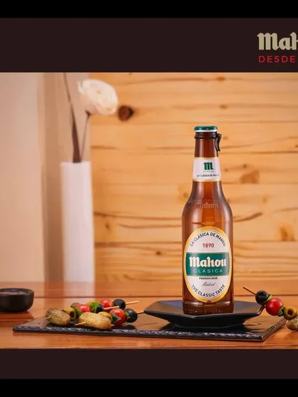 This image showcases a Mahou Clasica bottle with assorted skewers on a slate platter. The setup is designed to highlight the beer as a perfect companion for appetizers and social gatherings.