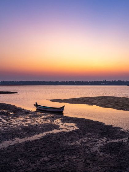 A lone boat is moored in the calm backwaters near Brahmavara as the sun sets. The image evokes the quiet, peaceful lifestyle of the coastal Karavali region.